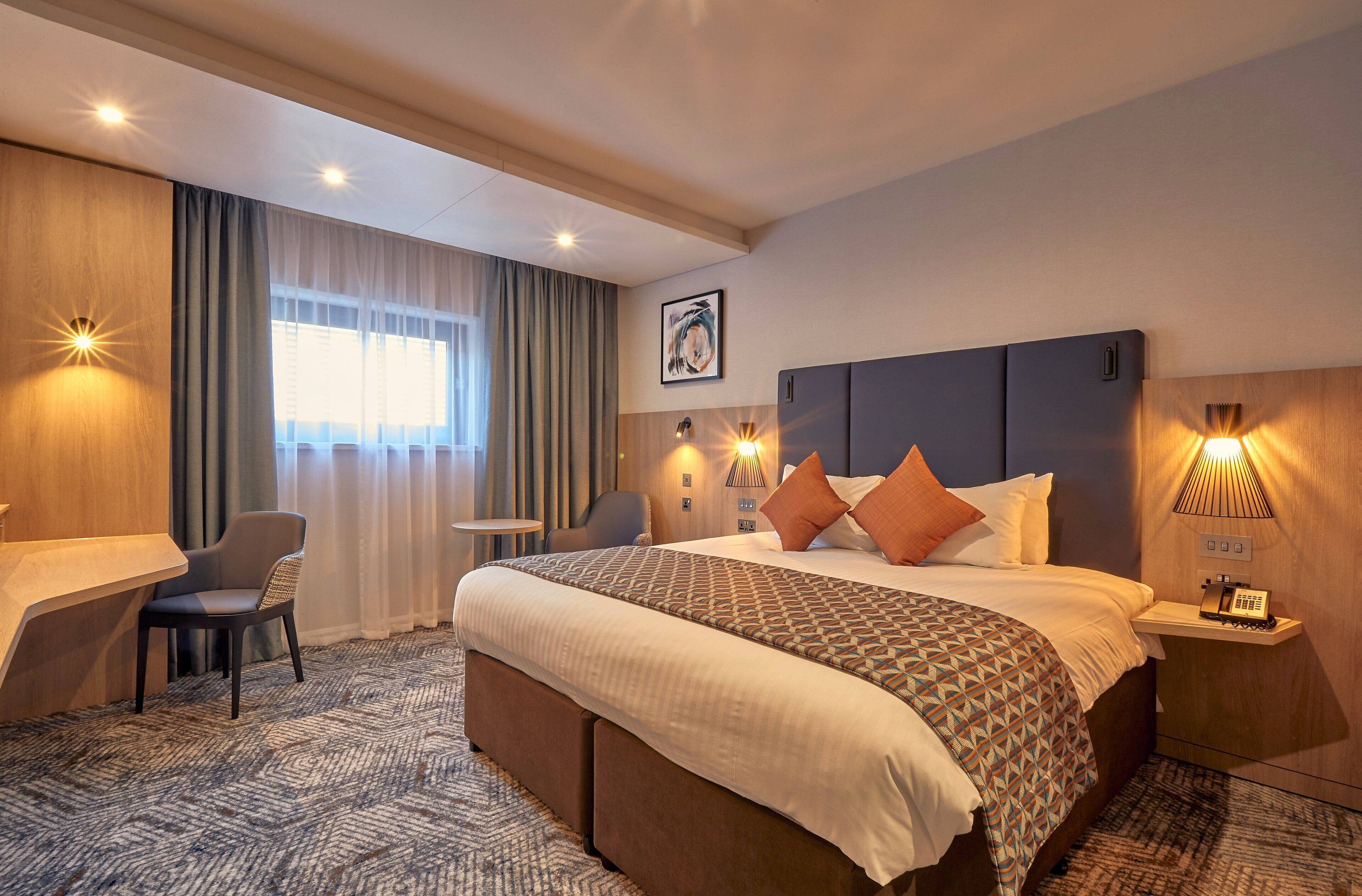 Habitación de hotel con cama, escritorio, silla, mesita de noche y lámpara de pared.