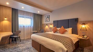 Habitación de hotel con cama, escritorio, silla, mesita de noche y lámpara de pared.