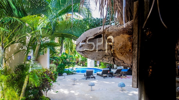 Zona de piscina con sillas de descanso, una formación rocosa con la palabra 'SPA' grabada y exuberante vegetación tropical.