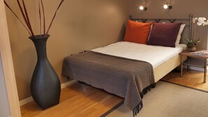Un dormitorio con una cama, una mesita de noche, un jarrón con ramas y una lámpara fijada a la pared.