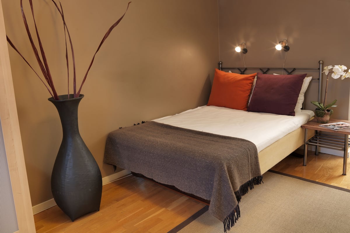 A bedroom with a bed, a nightstand, a vase with branches, and a wall-mounted light fixture.