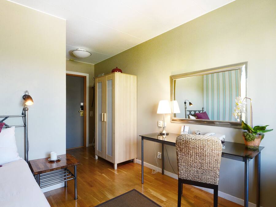 Habitación de hotel con cama, mesita de noche, lámpara, silla, escritorio, espejo y una planta en maceta.