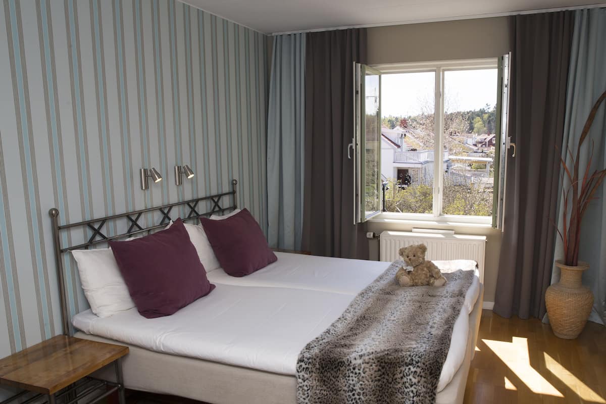 A bedroom with a bed, a bedside table, a window with curtains, and a view of the outside.