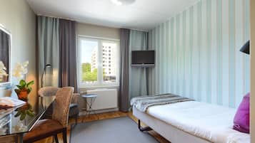 Ein Hotelzimmer mit einem Bett, einem Schreibtisch mit Stuhl, einem Fernseher, einem Fenster mit Vorhängen und Blick auf Gebäude.