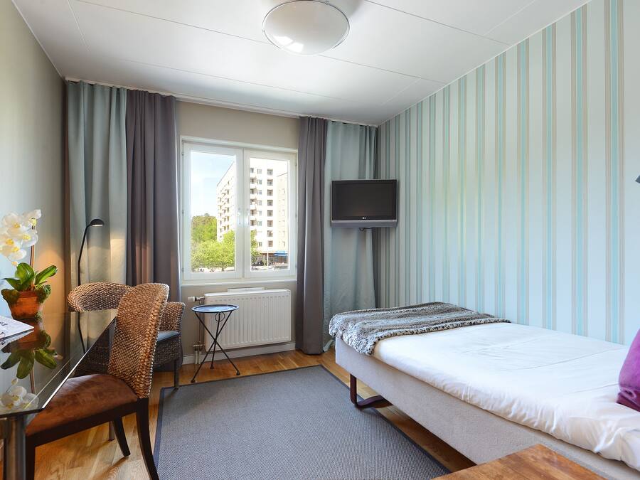 Ein Hotelzimmer mit einem Bett, einem Schreibtisch mit Stuhl, einem Fernseher, einem Fenster mit Vorhängen und Blick auf Gebäude.