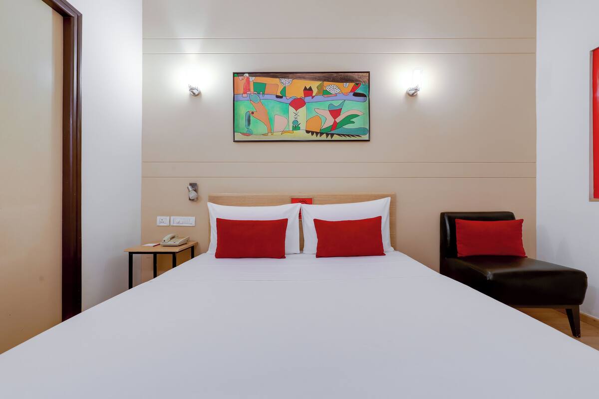 A hotel room with a bed, two red pillows, a nightstand with a phone, a painting on the wall, and a red chair.
