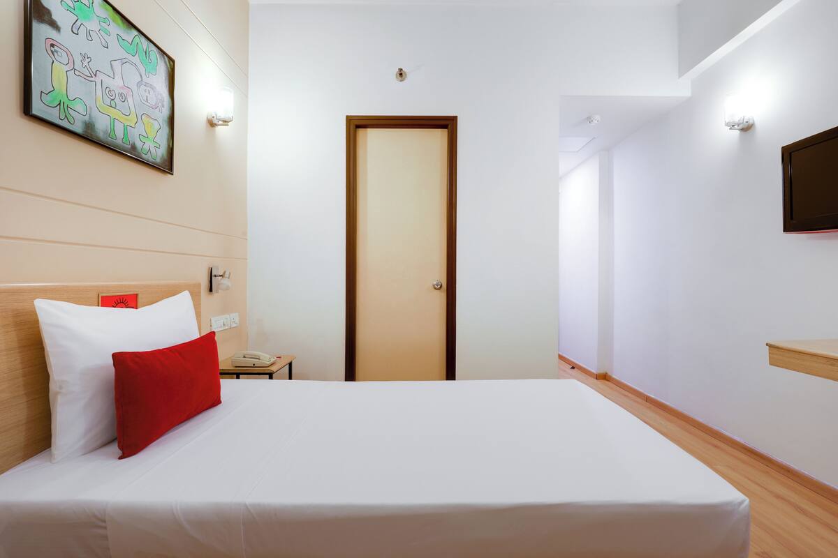 A hotel room with a bed, a wooden headboard, a red pillow, a white pillow, a wall-mounted TV, and a door.