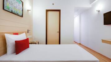 A hotel room with a bed, a wooden headboard, a red pillow, a white pillow, a wall-mounted TV, and a door.