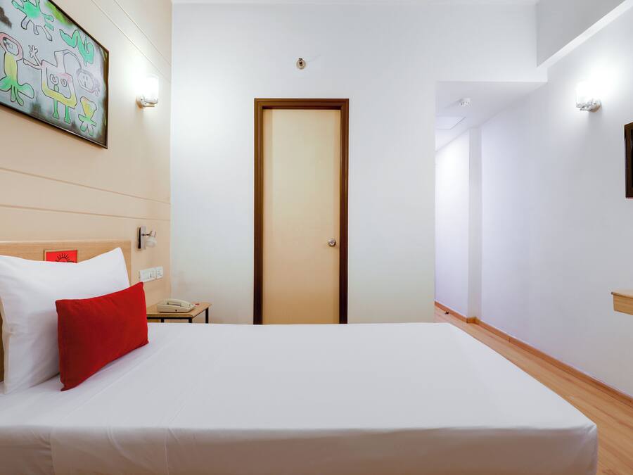 A hotel room with a bed, a wooden headboard, a red pillow, a white pillow, a wall-mounted TV, and a door.