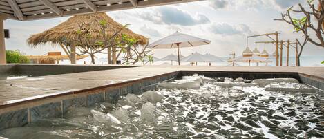 A hot tub area with a thatched roof structure, umbrellas, and a view of the sea.