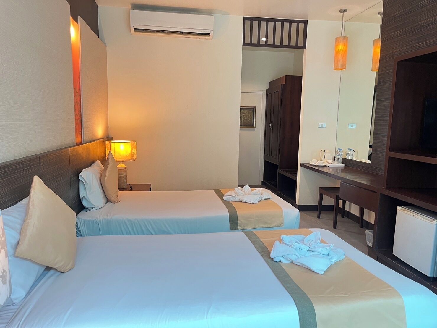 A hotel room with two beds, a desk, a chair, a mirror, a TV, and an air conditioning unit.