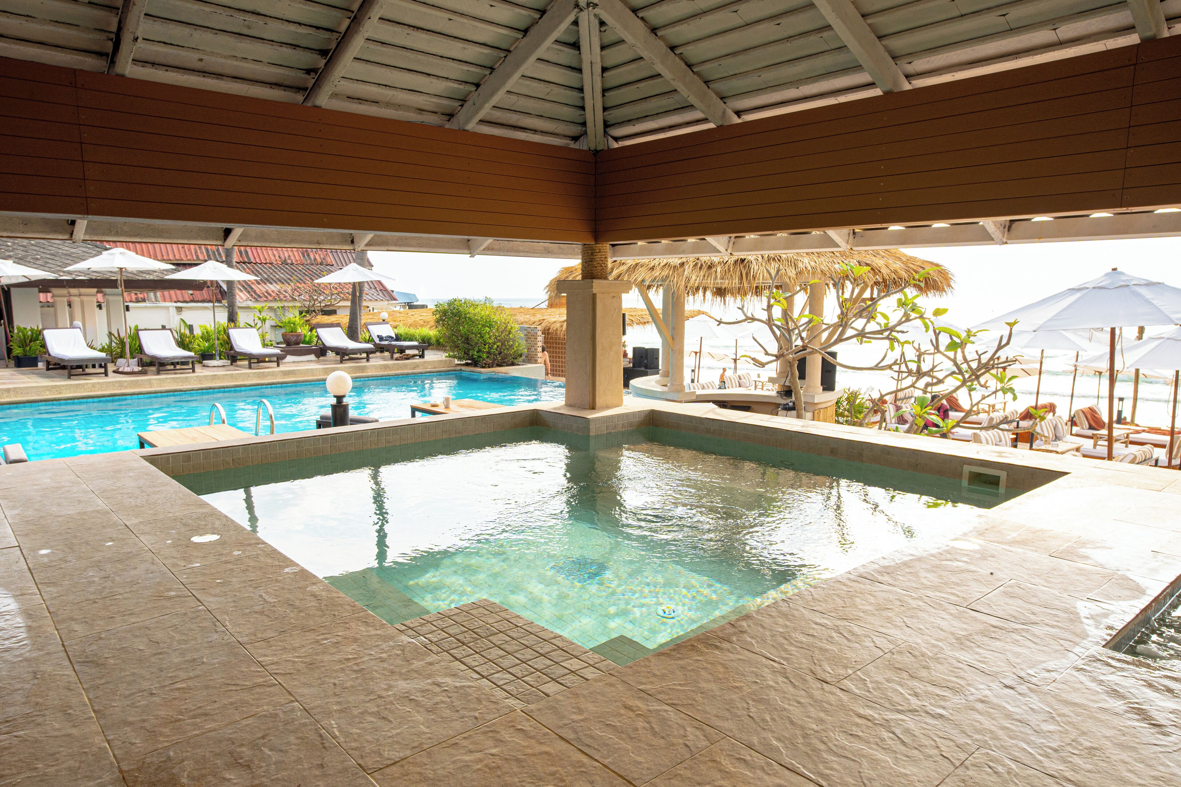 An indoor swimming pool with tiled flooring, lounge chairs, umbrellas, and a thatched roof structure.
