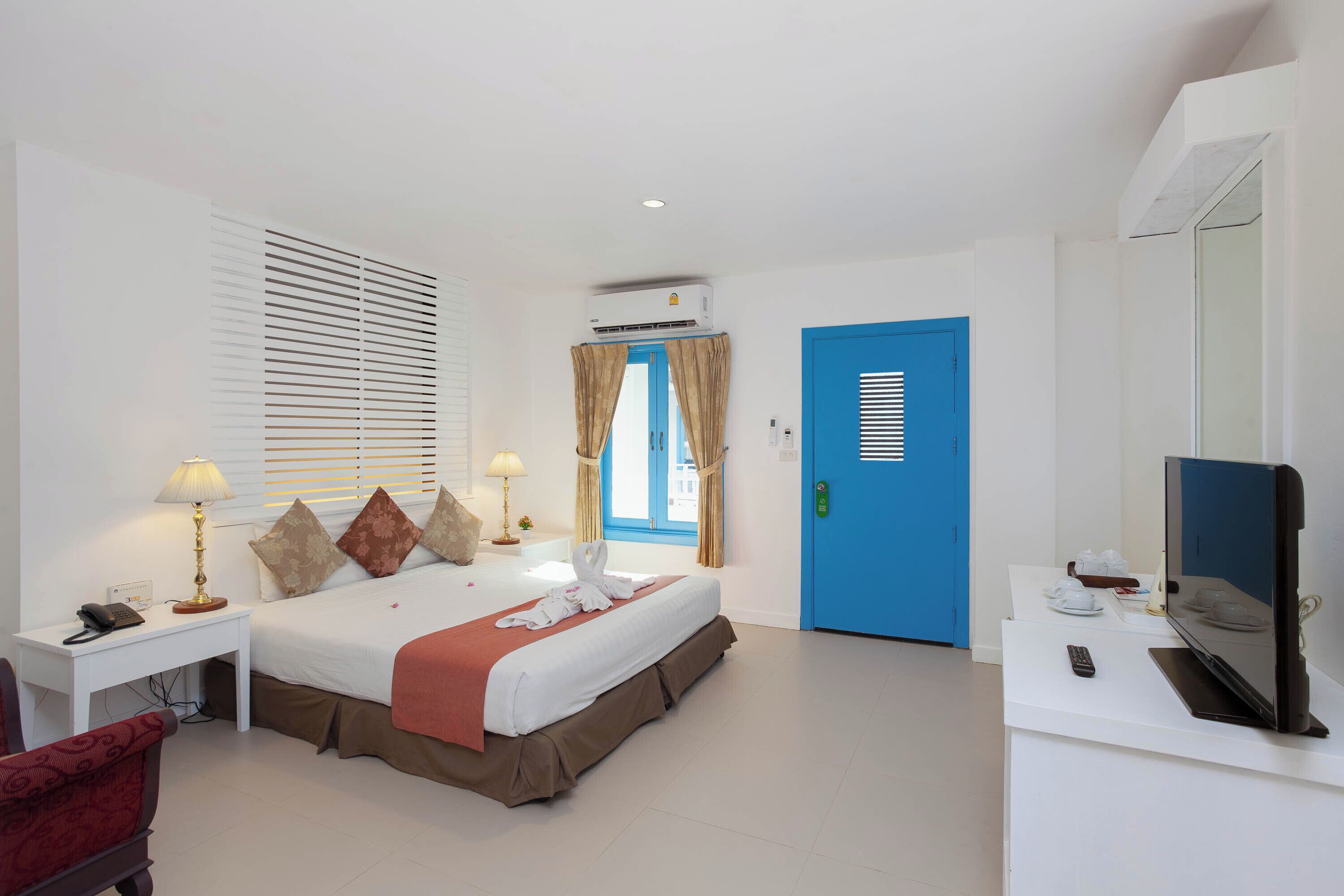 A hotel room with a bed, bedside lamps, a desk with a phone, a TV, and a blue door.