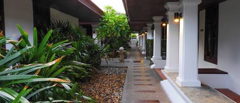 A covered corridor with white columns, a gravel path, and lush greenery.