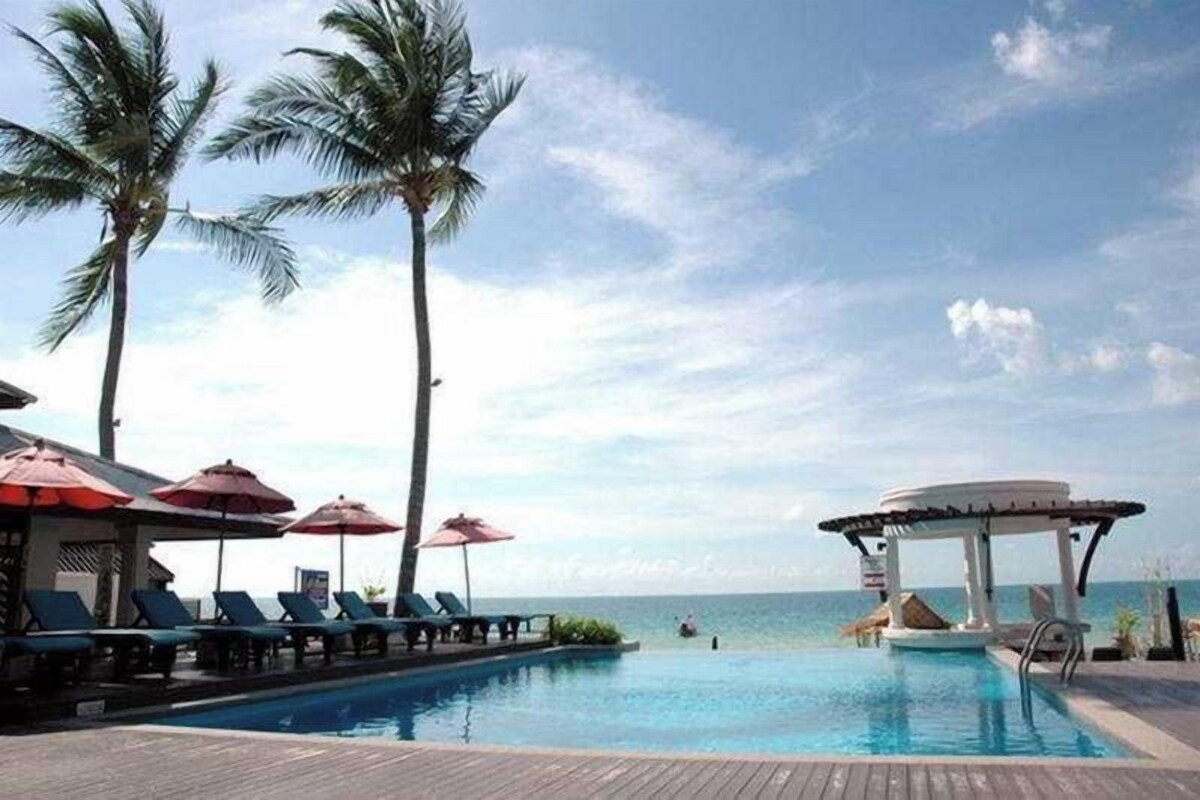 A poolside area with lounge chairs and umbrellas, palm trees, and a view of the sea.