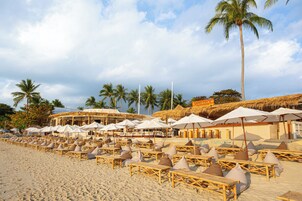 Een strand met rijen ligbedden en parasols, palmbomen en een rieten gebouw met de tekst 'SAND DECK'.
