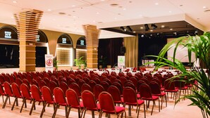 A conference room with rows of red chairs, a stage with a podium, and a large plant. - Guitart Central Park Aqua Resort (Lloret de Mar)
