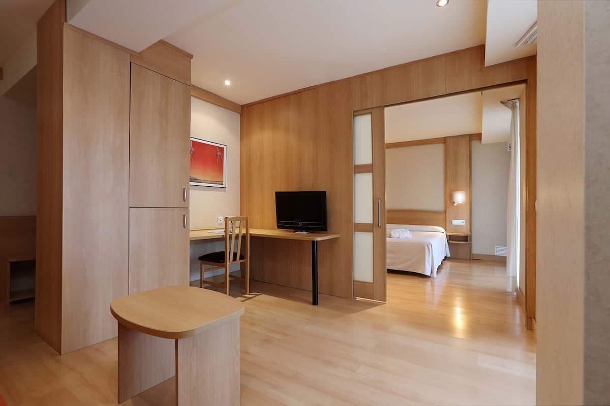 Ein modernes Hotelzimmer mit Holzboden, einem Schreibtisch mit Fernseher, einem Bett und einer Schiebetür zu einem weiteren Raum.