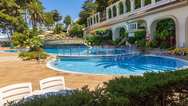 A pool area with a clear blue pool, white lounge chairs, and a building with arched windows and greenery. - Guitart Central Park Aqua Resort (Lloret de Mar)