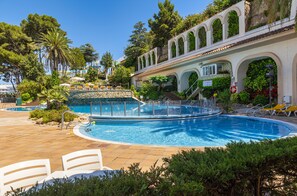 A pool area with a clear blue pool, white lounge chairs, and a building with arched windows and greenery. - Guitart Central Park Aqua Resort (Lloret de Mar)