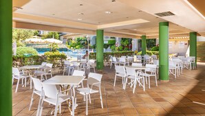 An outdoor cafe area with white tables and chairs, green pillars, and a view of a pool and greenery in the background. - Guitart Central Park Aqua Resort (Lloret de Mar)