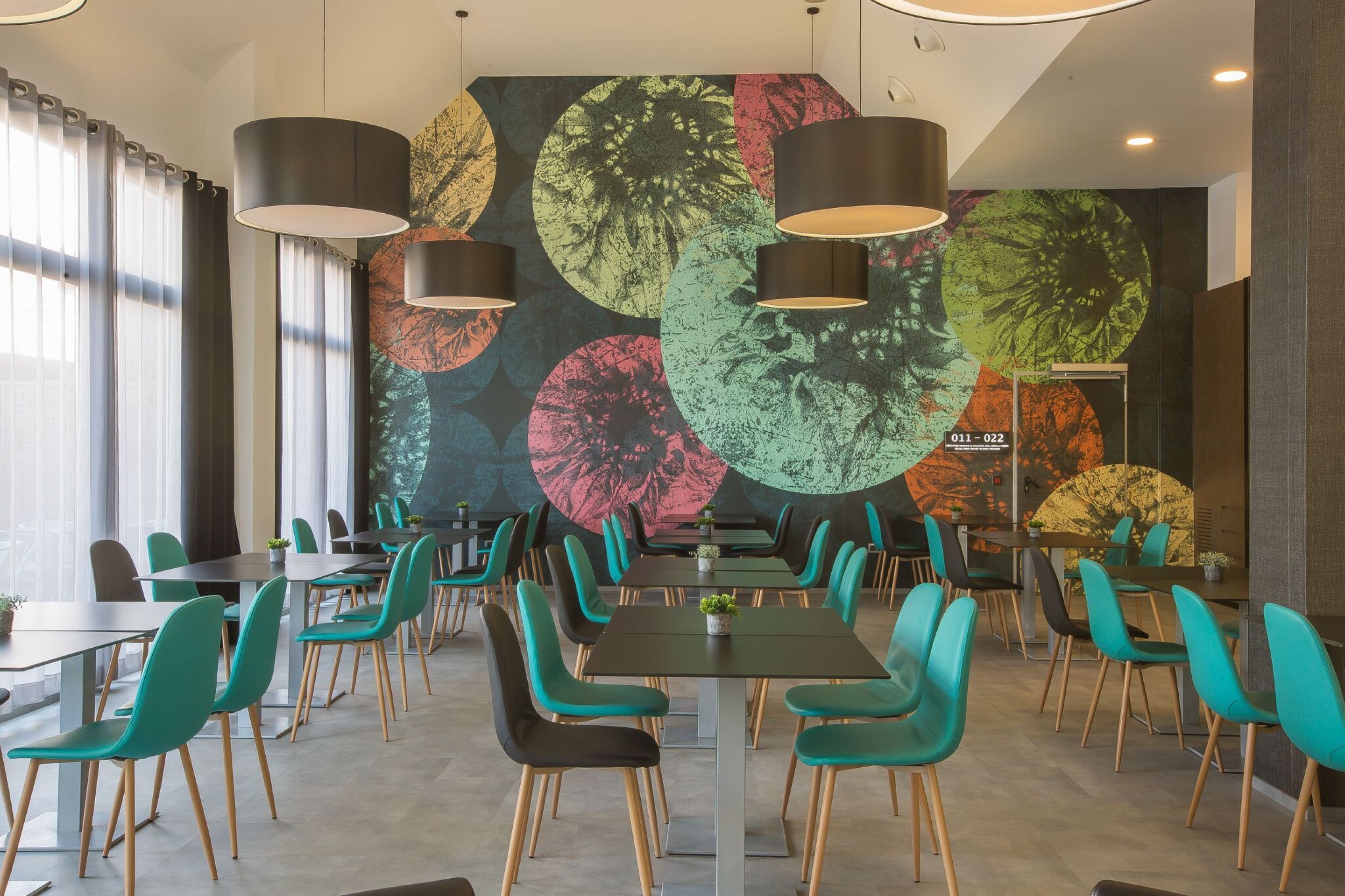 Et moderne spiseområde med teal farvede stole, sorte borde og et farverigt mural på væggen.