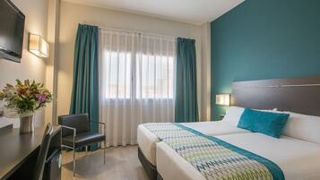 Una habitación de hotel con cama, escritorio, silla y ventana con cortinas.