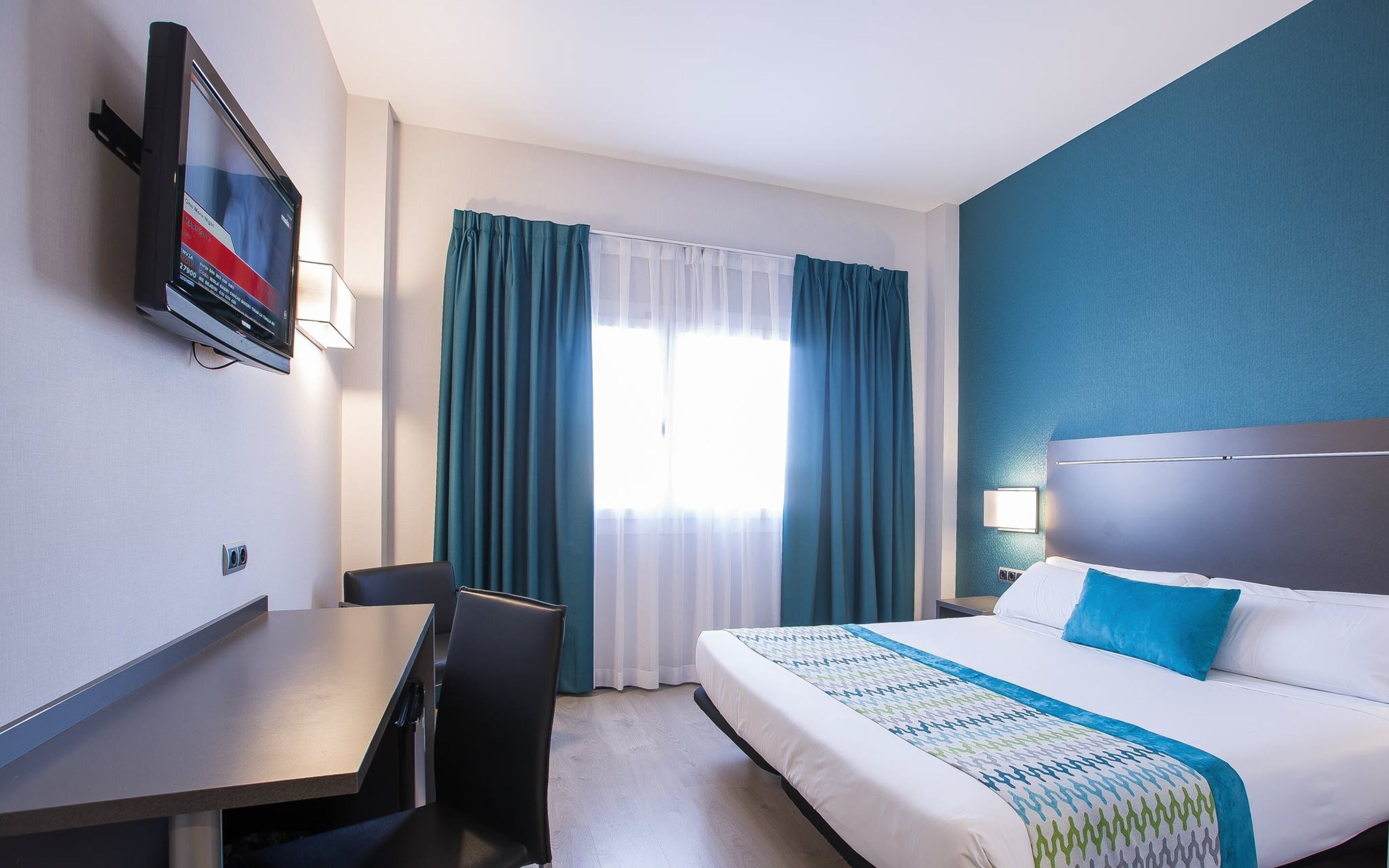 A hotel room with a bed, a desk, a chair, a TV mounted on the wall, and a window with curtains.