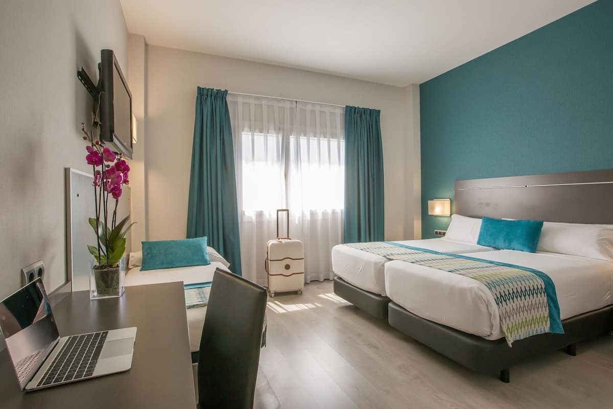 Een hotelkamer met een bed, een bureau met een laptop, een stoel, een koffer en een bloempot met plant.