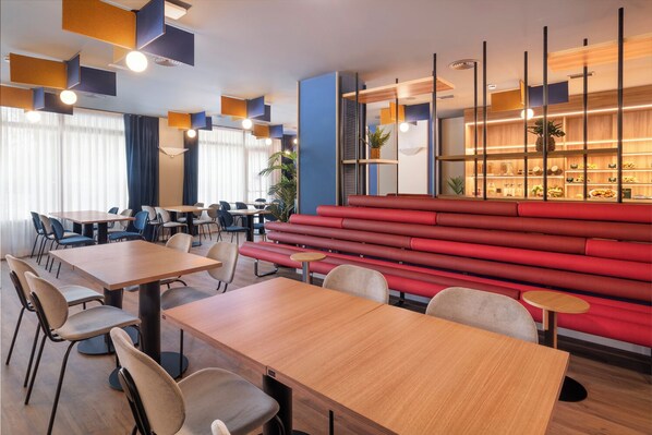 A modern dining area with wooden tables and chairs, a red bench seating area, and a wall with a built-in shelf displaying food items. - B&B Hotel Barcelona Rubí (Rubi)