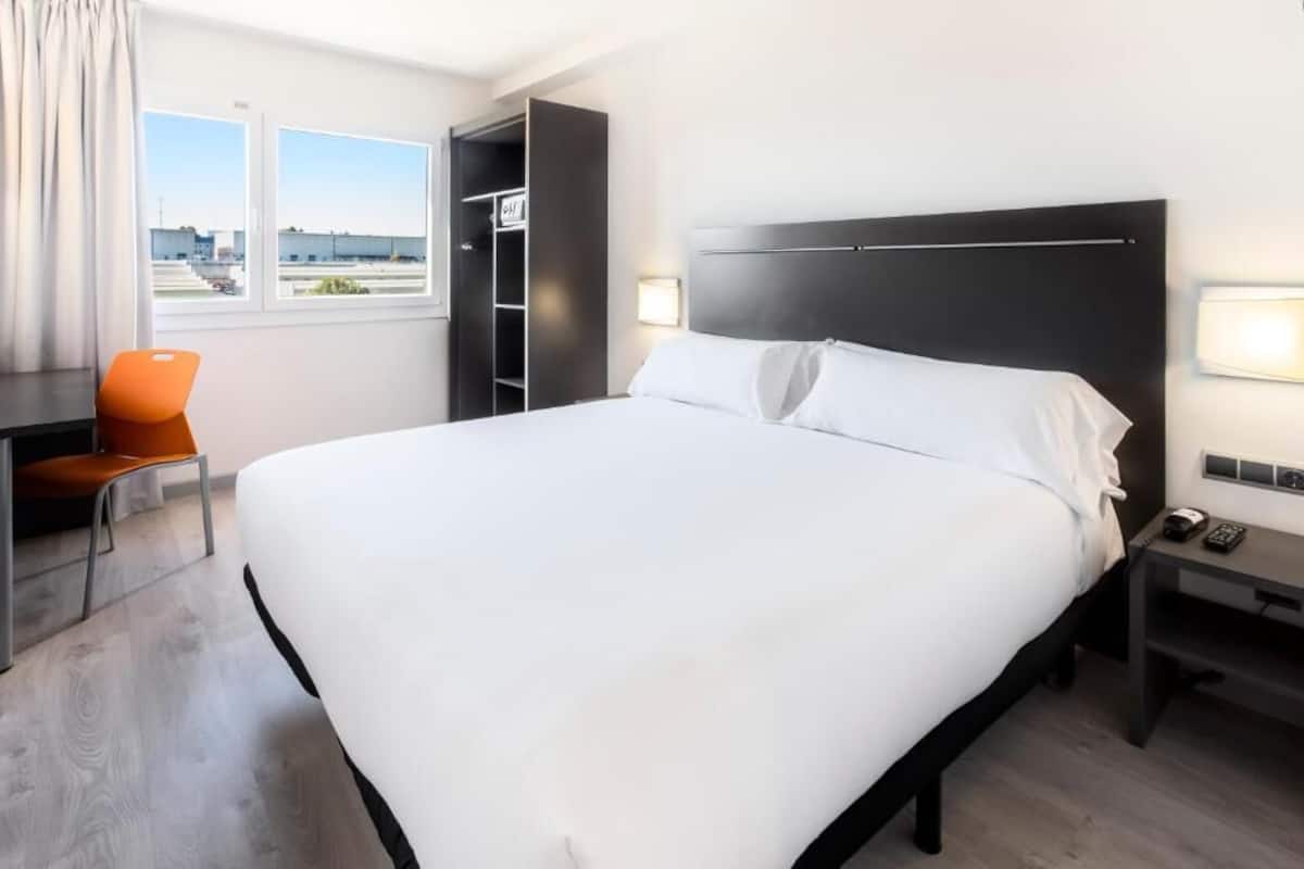 A hotel room with a large bed, a desk, a chair, a wardrobe, and a window with a view.