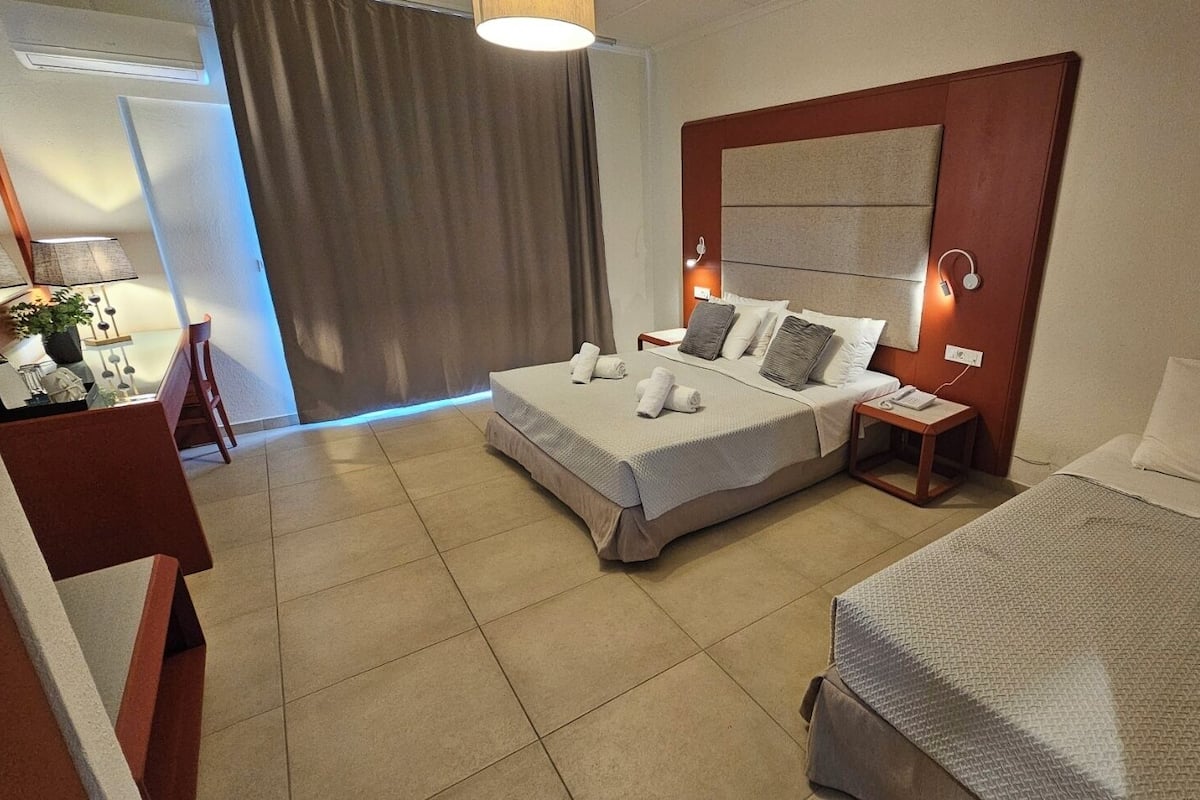Ein Hotelzimmer mit einem großen Bett, einem kleineren Bett, einem Schreibtisch mit Stuhl und einem durch einen Vorhang einsehbarer Badezimmer.