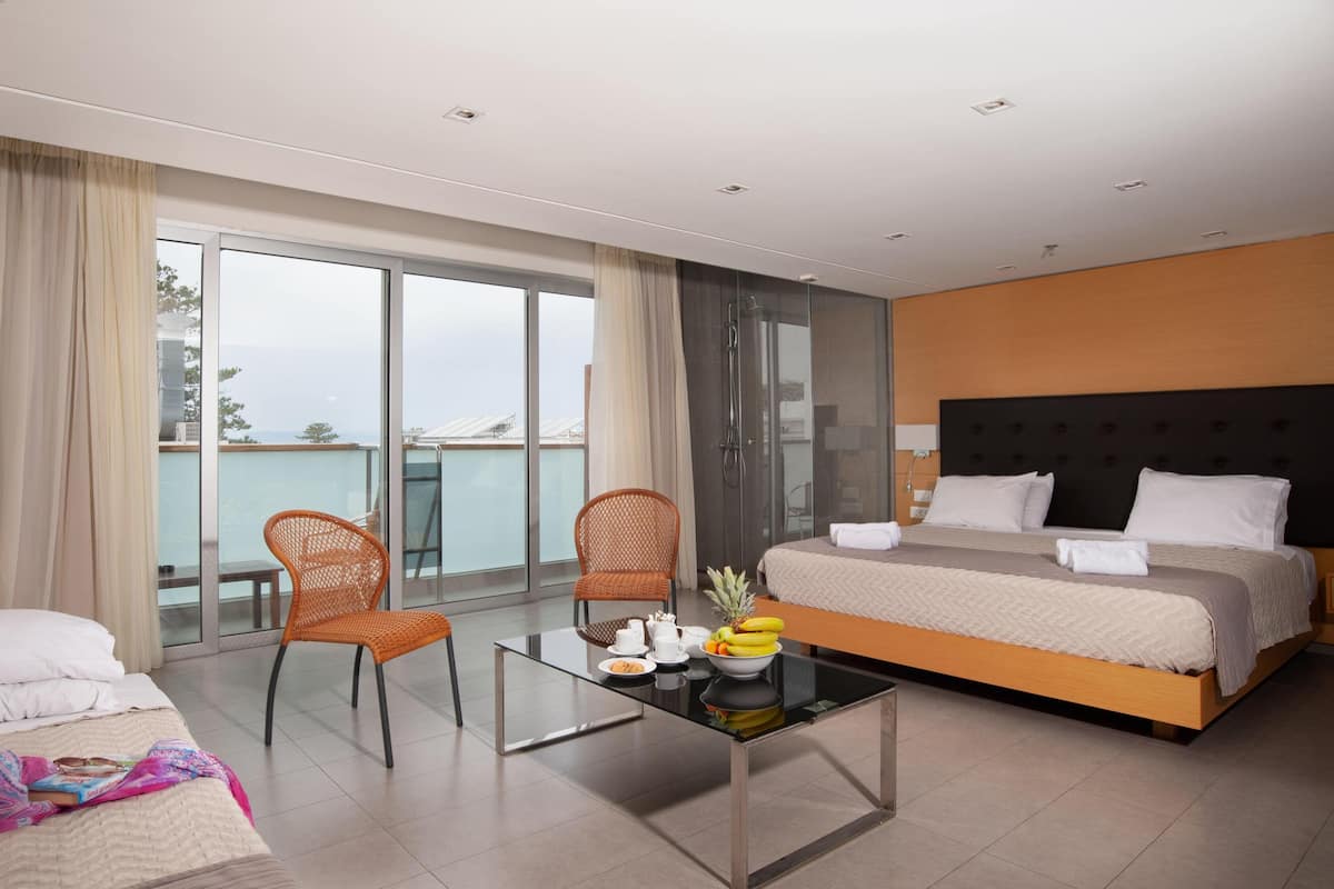 Ein modernes Hotelzimmer mit einem großen Bett, zwei Stühlen, einem Glastisch mit Frühstücksangeboten und einem Balkon mit Meerblick.