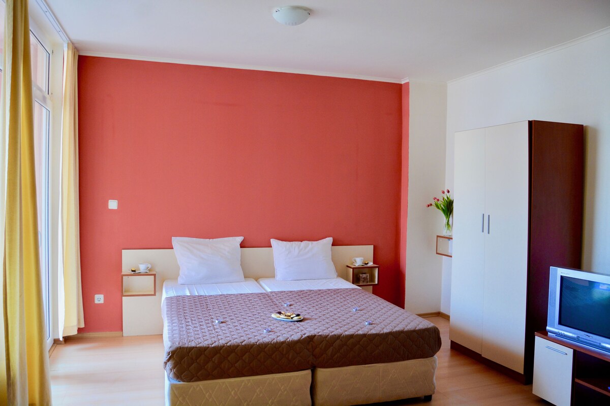 A double bed with a quilt, bedside tables with lamps, a TV stand, and a wardrobe.