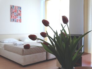 Una cama con ropa blanca y una pequeña planta en maceta con hojas verdes y flores rojas en un florero de vidrio transparente.