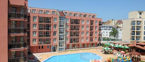A multi-story hotel with a swimming pool, sun loungers, and a children's play area.