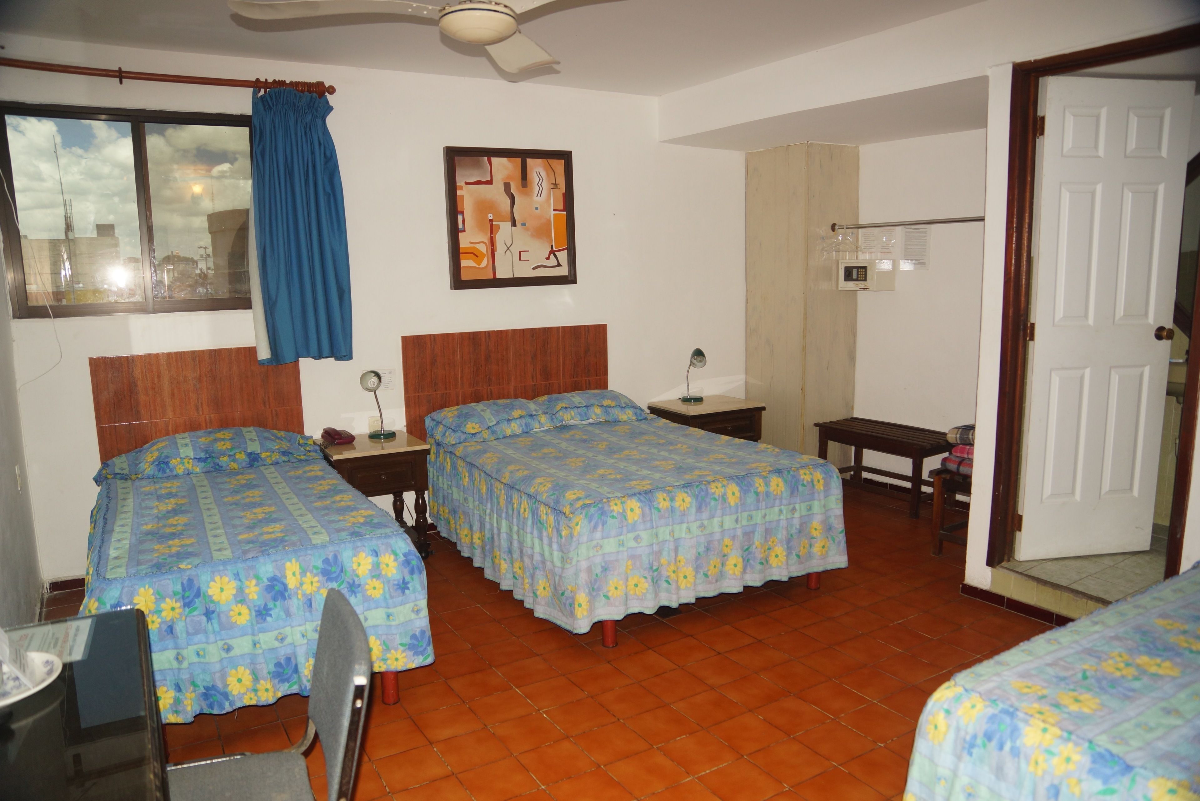 A room with two beds, a desk, a chair, a ceiling fan, a window with curtains, and a wall-mounted artwork.