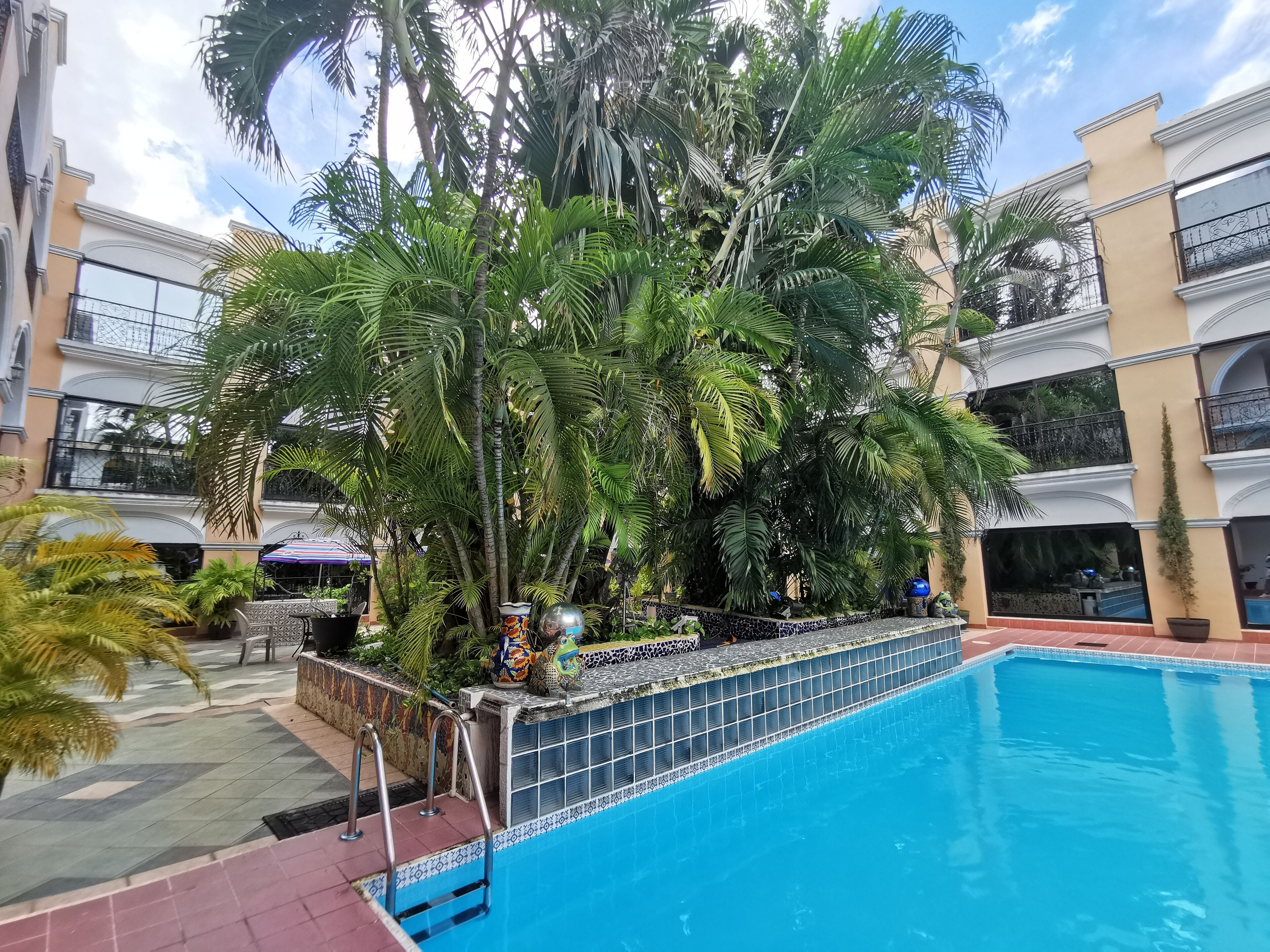 A swimming pool surrounded by lush tropical plants and a multi-story building with balconies.