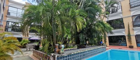 A swimming pool surrounded by lush tropical plants and a multi-story building with balconies.