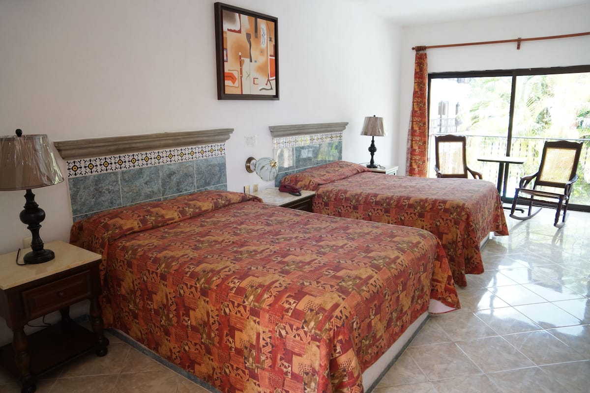 A hotel room with two beds, a balcony, and a view of greenery.