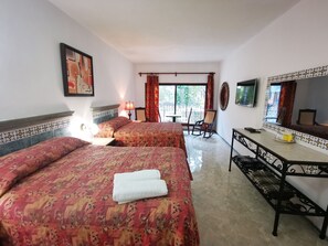 A hotel room with two beds, a desk, a chair, a TV, and a balcony with curtains.