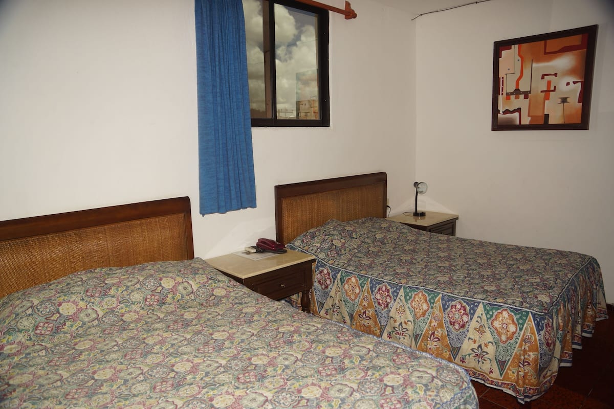 A room with two single beds, a window with blue curtains, a bedside table with a lamp, and a wall-mounted abstract painting.