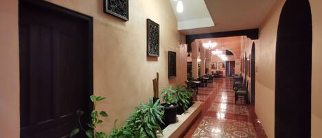 A corridor with a tiled floor, potted plants, framed artwork, and a seating area with tables and chairs.