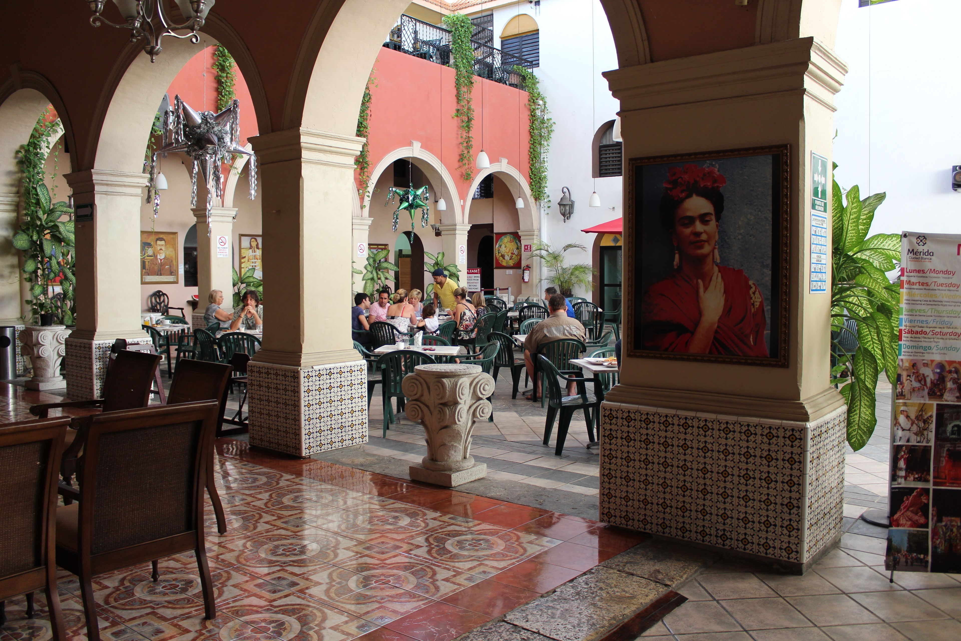 An indoor courtyard with tiled flooring, green chairs, and tables. There is a large framed portrait on a pedestal and a sign with a list of days and times.