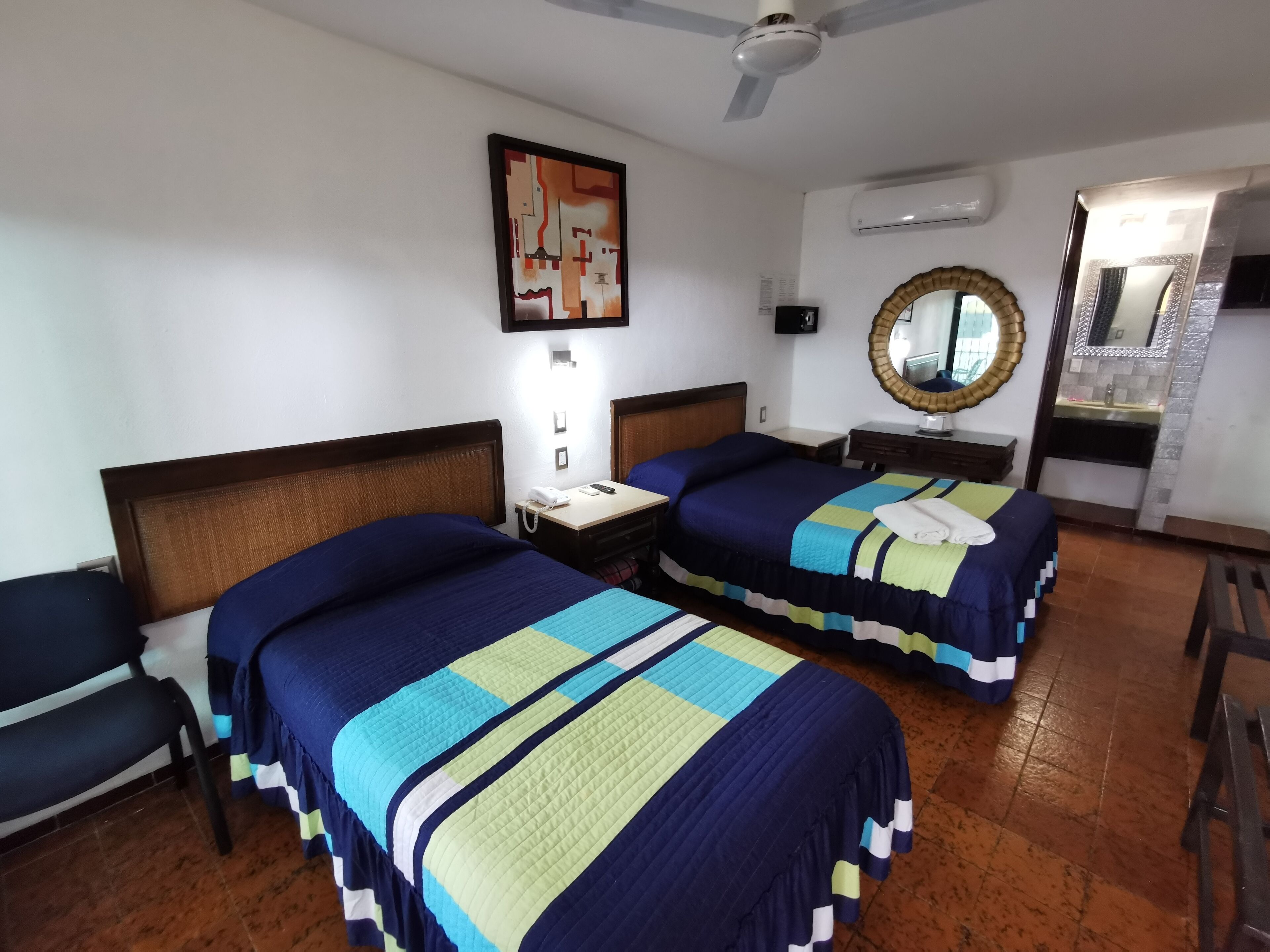 Habitación de hotel con dos camas, un ventilador de techo, un equipo de aire acondicionado, un espejo y una mesita con un teléfono y una lámpara.
