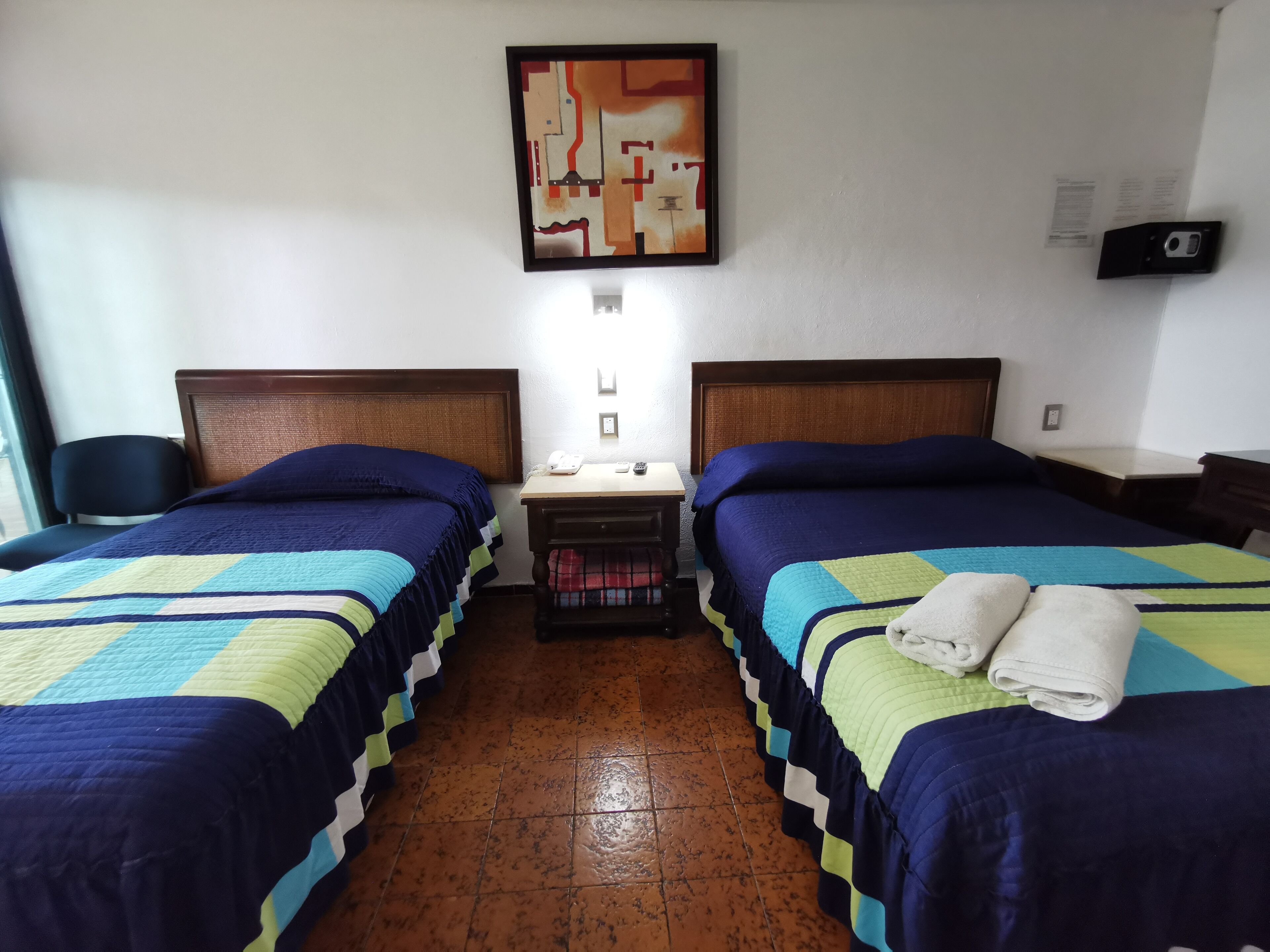 A hotel room with two beds, each with a blue and green quilt, a nightstand with a phone and a lamp, and a wall-mounted painting.