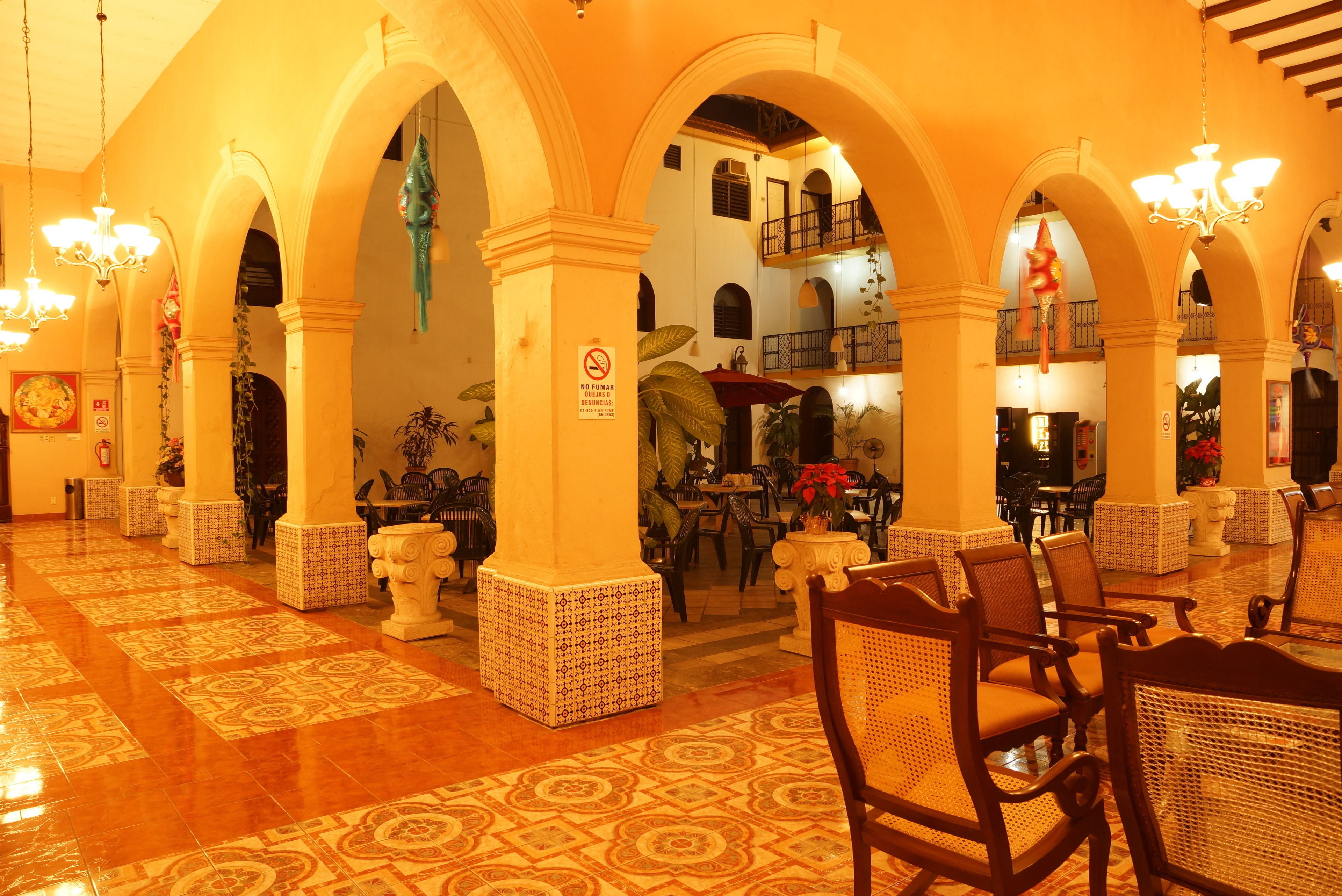 A lobby with a tiled floor, wooden chairs, and tables. There are arches and columns, and a sign prohibiting smoking.