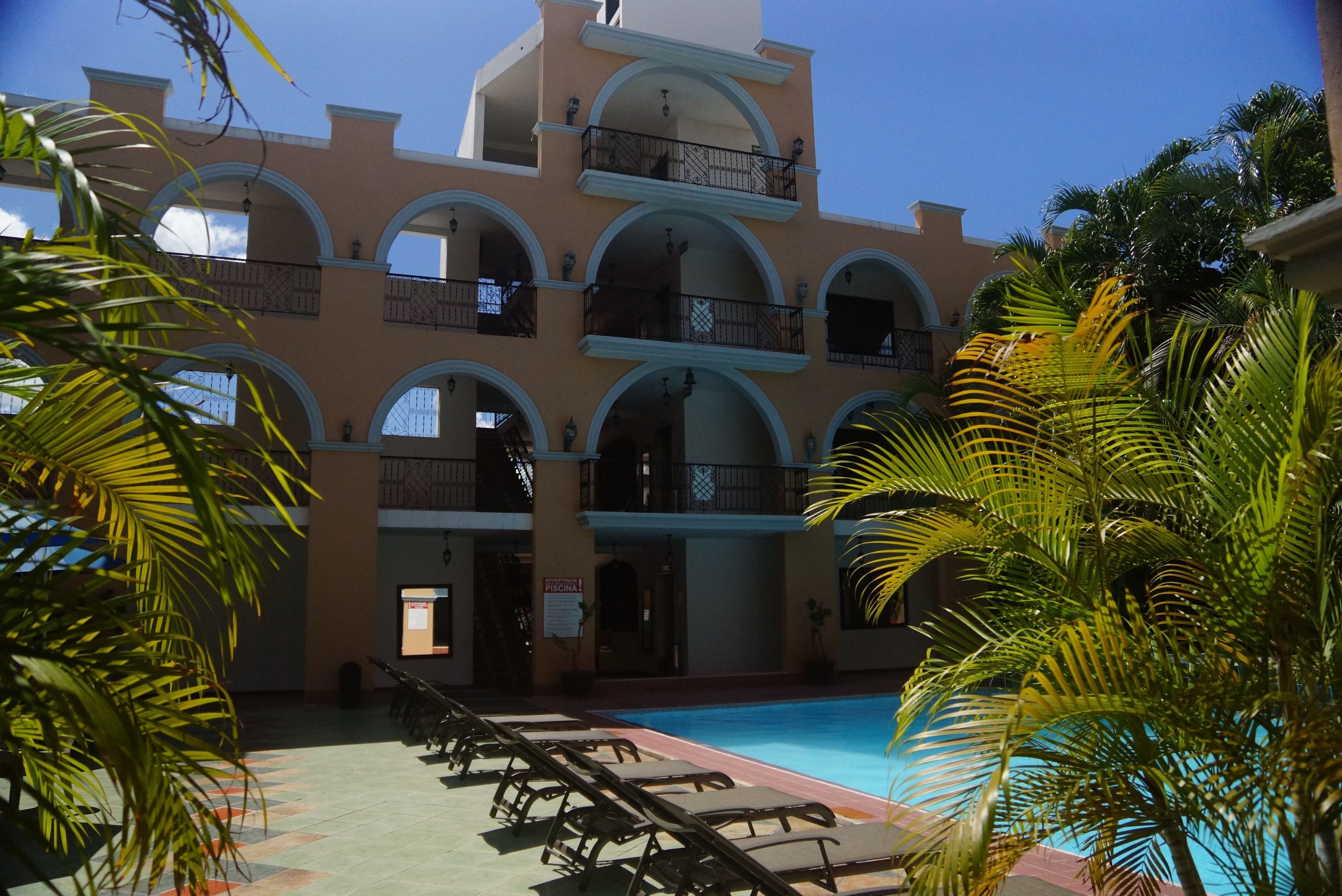 A multi-story building with a pool area, featuring a series of balconies and a clear blue sky.