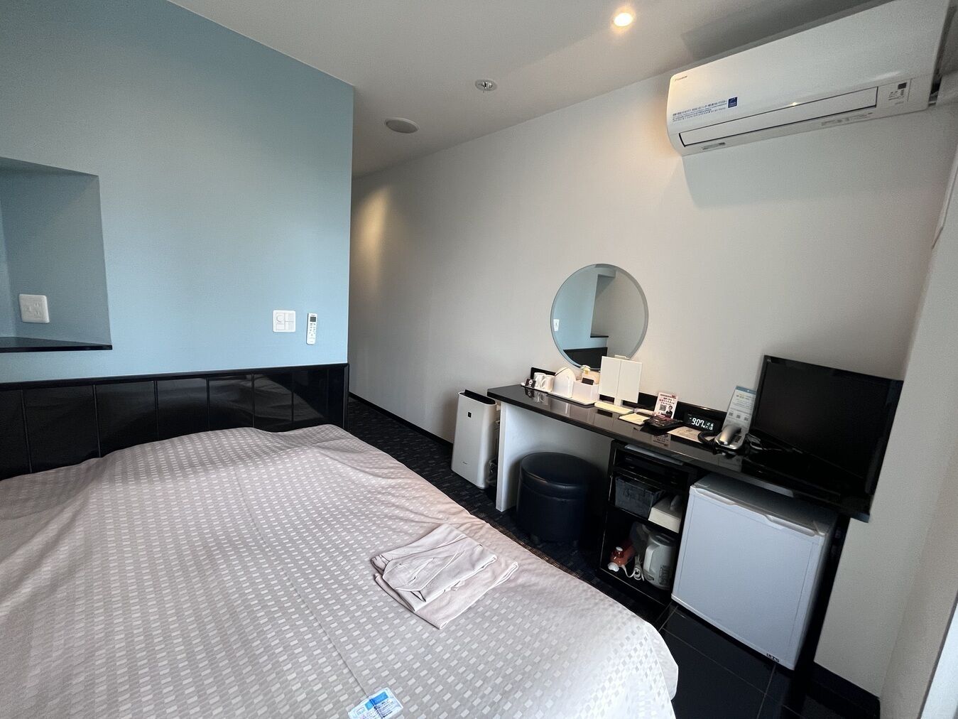 A hotel room with a bed, a desk with a computer, a mirror, an air conditioner, and a small refrigerator.