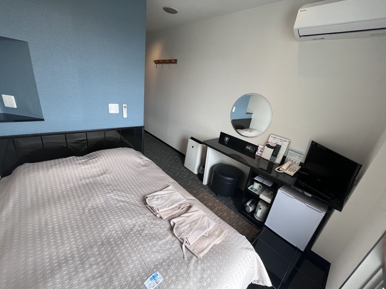 A hotel room with a bed, a desk with a computer, a mirror, and a small refrigerator.
